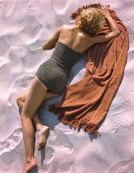 Photographie vintage colorisée d'un mannequin femme en tenue élégante des années 1940 (grand chapeau de paille, jupe rayée) debout sur une vaste plage de sable face à l'océan.