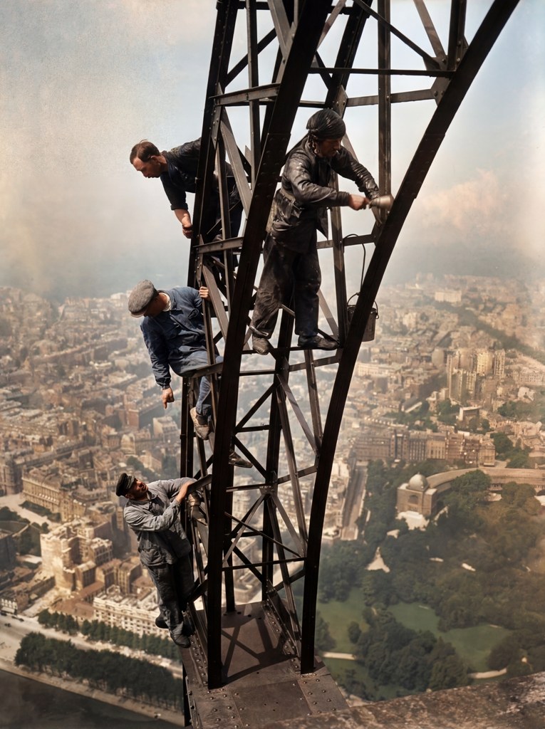 Photographie historique colorisée des ouvriers peintres travaillant sur la structure en fer de la Tour Eiffel, surplombant le Champ-de-Mars et Paris.