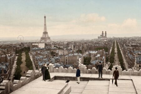Vue panoramique colorisée de Paris en 1900 depuis un toit. On aperçoit la Tour Eiffel, l'ancien Trocadéro et des Parisiens en costumes d'époque (hauts-de-forme et robes longues).