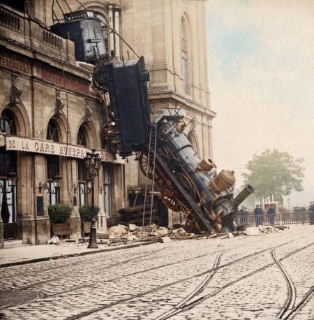 Photographie historique colorisée de la locomotive à vapeur gisant sur le trottoir après avoir traversé la façade de la Gare Montparnasse à Paris en 1895.