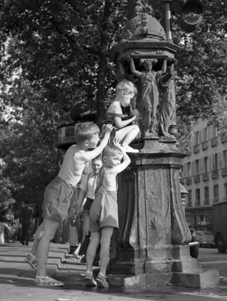 Photographie d'époque colorisée montrant trois petits garçons jouant et se rafraîchissant autour d'une fontaine Wallace dans une rue de Paris. Scène de vie nostalgique.