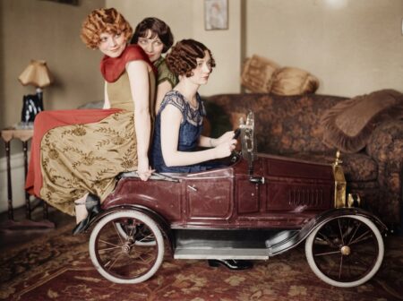 Photographie colorisée des Années Folles montrant trois jeunes femmes élégantes en robes d'époque posant avec une voiture à pédales miniature dans un salon.
