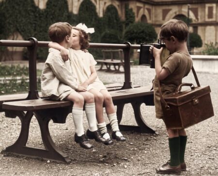 Photographie vintage colorisée de 1930 montrant un jeune garçon photographiant deux autres enfants en train de s'embrasser sur un banc dans un parc.