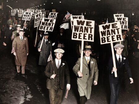 Photographie historique colorisée montrant une foule d'hommes en costume manifestant la nuit contre la Prohibition aux États-Unis, brandissant des panneaux "WE WANT BEER".