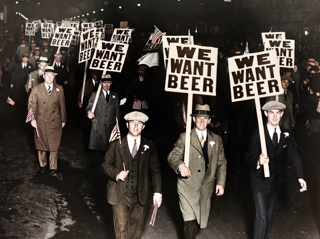 Photographie historique colorisée montrant une foule d'hommes en costume manifestant la nuit contre la Prohibition aux États-Unis, brandissant des panneaux "WE WANT BEER".