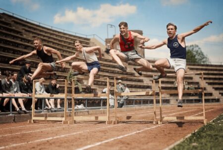 Photographie historique colorisée montrant quatre jeunes athlètes franchissant des haies en bois sur une piste d'athlétisme dans un stade des années 30.