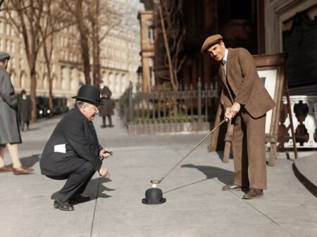 Photographie historique colorisée de 1910 montrant deux hommes en costume dans une rue. L'un utilise un chapeau melon pour poser une balle de golf tandis que l'autre se prépare à frapper avec son club.
