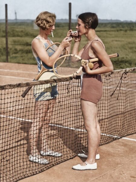 Photographie historique colorisée montrant deux jeunes femmes en tenue de sport des années 1920 s'accordant une pause au filet d'un court de tennis pour allumer une cigarette, leurs raquettes en bois sous le bras.
