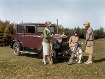Photographie historique colorisée des années 1920 montrant trois femmes élégantes en tenue de golf discutant près d'une voiture d'époque bordeaux dans un paysage de campagne.