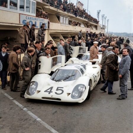 Photographie historique colorisée montrant deux voitures de course Porsche 908 blanches dans les stands du Mans en 1969, entourées de mécaniciens et de spectateurs.