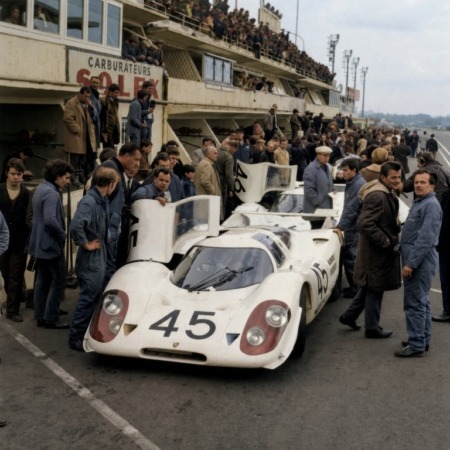 Photographie historique colorisée montrant deux voitures de course Porsche 908 blanches dans les stands du Mans en 1969, entourées de mécaniciens et de spectateurs.