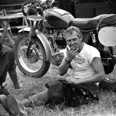 Photographie noir et blanc de l'acteur Steve McQueen souriant avec une cigarette, assis dans l'herbe devant une moto Triumph.
