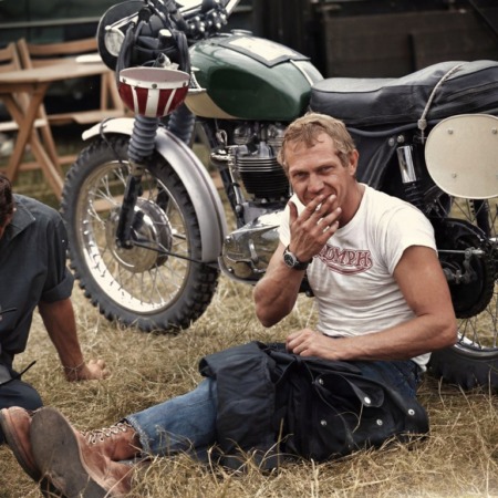 Photographie noir et blanc de l'acteur Steve McQueen souriant avec une cigarette, assis dans l'herbe devant une moto Triumph.