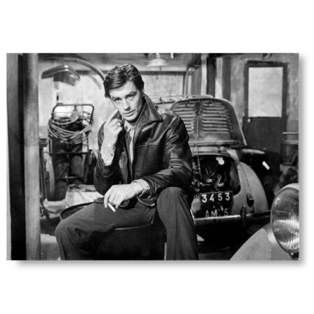 Photographie de cinéma en noir et blanc d'Alain Delon en blouson de cuir, assis devant une voiture ancienne dans un garage, tirée du film Mélodie en sous-sol.
