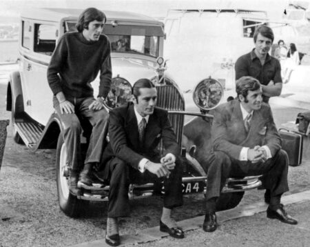 Photographie en noir et blanc d'Alain Delon et Jean-Paul Belmondo assis sur le pare-chocs d'une voiture d'époque, accompagnés de leurs frères debout derrière eux, sur les coulisses du tournage du film Borsalino.