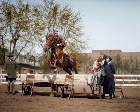 Affiche Cheval Saut Vintage : photographie d’art colorisée vers 1930 montrant un cavalier et son cheval sautant au-dessus d'une table dressée où sont assises deux femmes avec un chien Collie, lors d'une démonstration équestre.