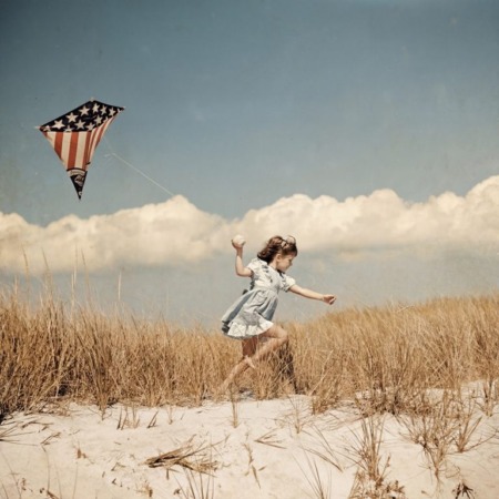 Affiche Cerf-Volant Vintage Plage : photographie d'art colorisée des années 50 montrant une petite fille courant dans les dunes avec un cerf-volant aux couleurs du drapeau américain sous un ciel d'été parsemé de nuages.
