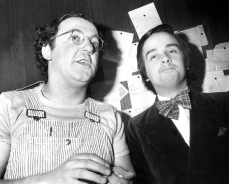 Affiche Coluche Le Luron : photographie d'art en noir et blanc de 1975 montrant Coluche en salopette à rayures et Thierry Le Luron en smoking avec un nœud papillon, capturés en coulisses.