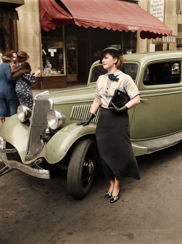 Affiche Mode Automobile Vintage Élégance : photographie d'art colorisée vers 1935 montrant une femme élégante en chapeau et gants posant à côté d'une Ford V8 verte des années 1930 dans une rue urbaine avec un auvent rouge.