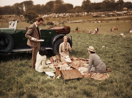Affiche Pique-Nique Élégance Campagne : photographie d’art colorisée montrant un groupe de quatre personnes chic en tenue des années 1920 (homme en tweed, femmes en chapeaux cloche) pique-niquant sur une colline à côté d’un grand cabriolet vert vintage.