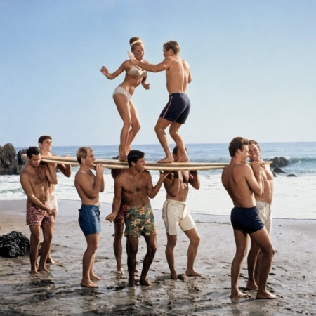Affiche Surf Vintage Plage : photographie d'art des années 60 montrant un groupe de jeunes surfeurs portant une planche sur leurs épaules, avec un couple dansant en équilibre au-dessus d'eux sur le sable.
