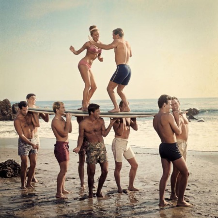 Affiche Surf Vintage Plage : photographie d'art des années 60 montrant un groupe de jeunes surfeurs portant une planche sur leurs épaules, avec un couple dansant en équilibre au-dessus d'eux sur le sable.