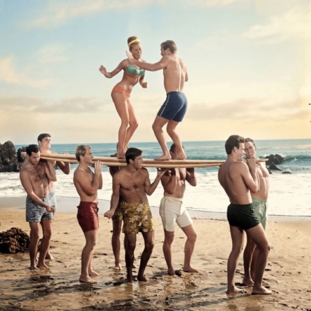 Affiche Surf Vintage Plage : photographie d'art des années 60 montrant un groupe de jeunes surfeurs portant une planche sur leurs épaules, avec un couple dansant en équilibre au-dessus d'eux sur le sable.