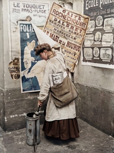 Affiche vintage en couleur d'une jeune femme collant des affiches dans une rue de Paris à la Belle Époque, devant un mur de réclames anciennes.